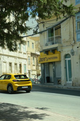 A yellow car driving past Jeff's Pastizzeria on a sunny day in a Mediterranean town.