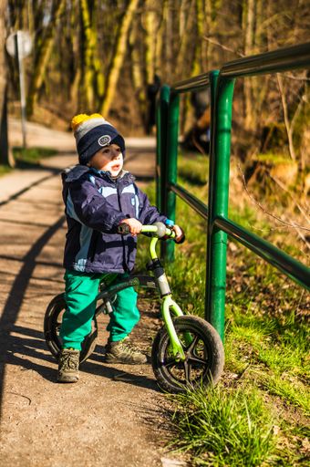 Small young boy sitting on a small balance bike by a green barrier in the Debiec forest.