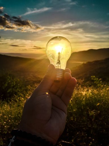 A hand holding a light bulb with reflection of a sun during sunset on mountains