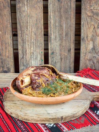 A traditional dish of roasted pork knuckle served on a clay plate with sauerkraut and herbs on a rustic wooden background.