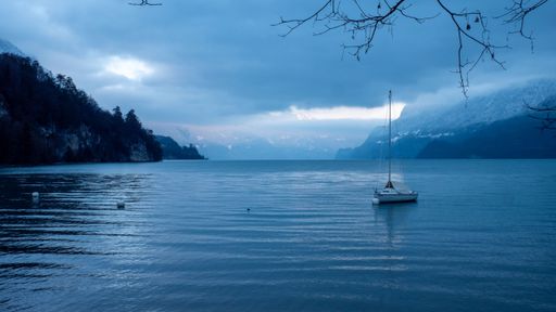 A serene winter lake scene with a sailboat and snow-covered mountains under a cloudy sky