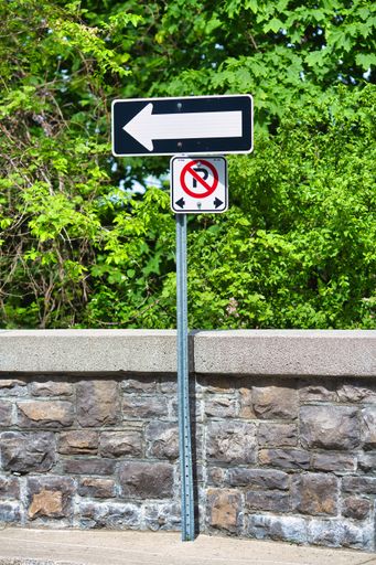 A metal pole supporting two road signs against a background of green foliage and a stone wall. The upper sign is a directional arrow indicating a left