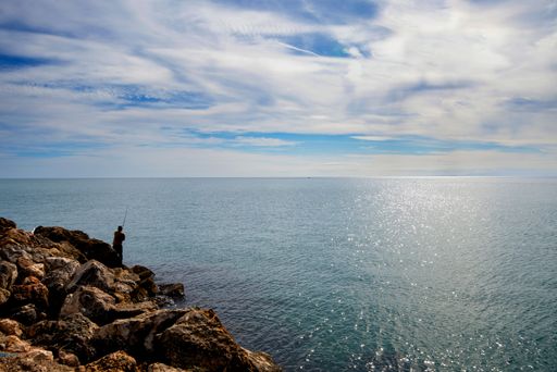 A lone fisherman standing on rocks by the sea under a partly cloudy sky on a sunny day