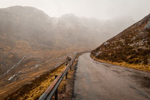 A winding mountain road with a guardrail on a foggy day, surrounded by rugged terrain and misty hills of Scotland.