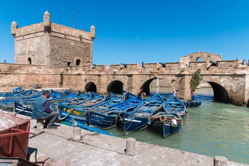 A summer day at the harbor in Essaouira, Morocco with small blue boats parked next to an arch bridge