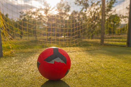 A red soccer ball on a green field in front of a goal net during sunset