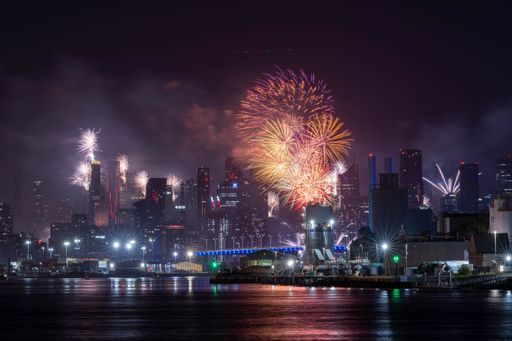 A vibrant cityscape at night with colorful fireworks lighting up the sky over a river.