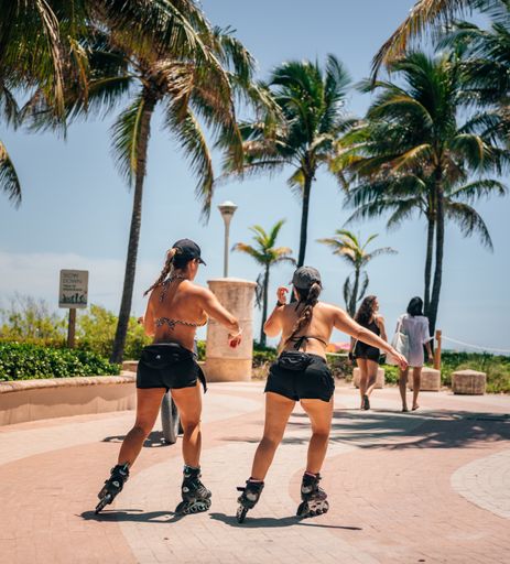 A vertical shot of two women in black shorts and rollerblades skating in the street with palm trees.