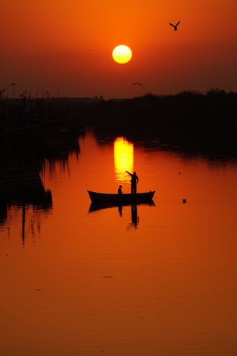 A vertical silhouette shot of two people on a boat in the lake, during a bright orange sunset