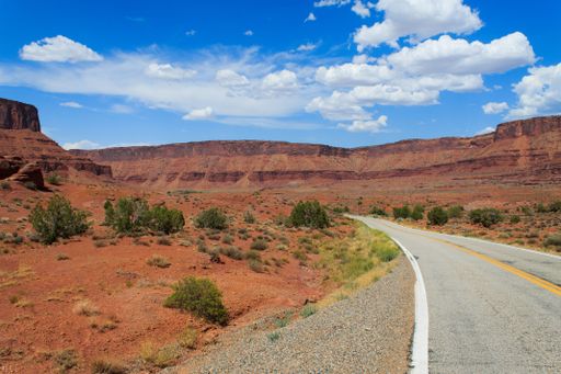 A scenic road winding through the red rock desert landscape under a bright blue sky with scattered clouds.