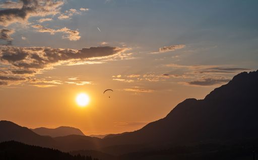 Paraglider flying over a scenic mountain landscape during sunset with a vibrant sky.