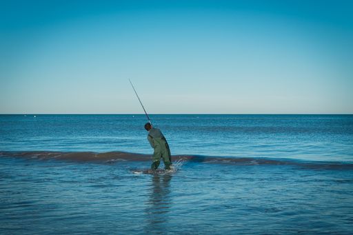 A man fishing intently in the blue sea as the tides wash over his knees at sunset under a clear sky