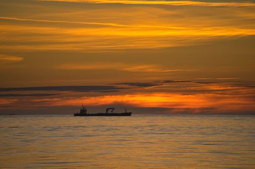A silhouette of a cargo ship against a vibrant orange sunset over calm ocean waters
