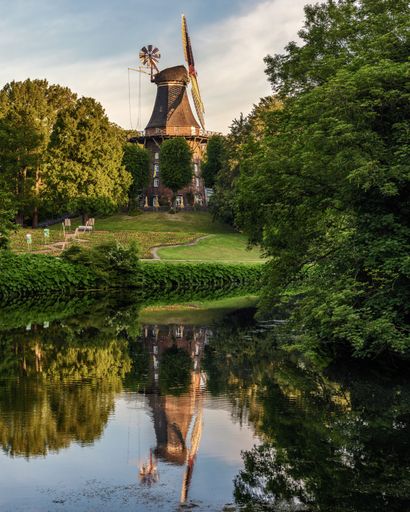 A picturesque windmill surrounded by lush greenery and reflected in a calm pond in Bremen, Germany.