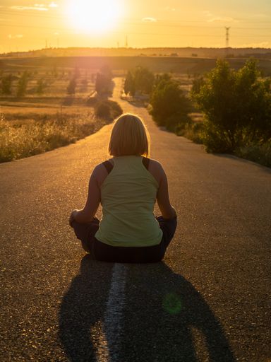 A vertical shot of a young female sitting on a road with her back and facing the sunset