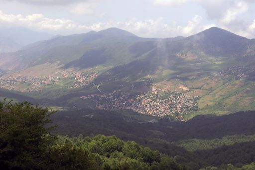 A landscape of a town surrounded by hills covered in greenery under a cloudy sky and sunlight in the countryside