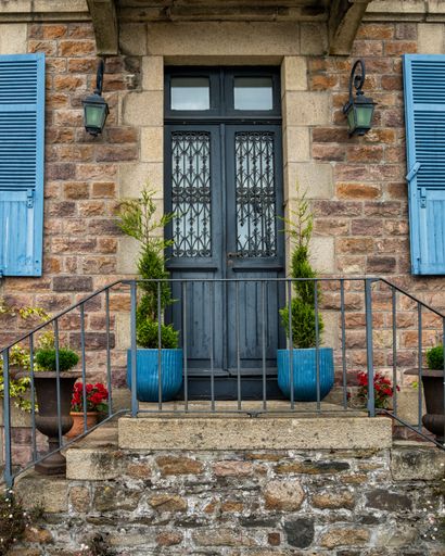 The charming entrance of a rustic stone house with blue shutters, potted plants, and vintage lanterns.