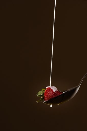 A vertical shot of a strawberry with milk on a spoon against a dark brown background
