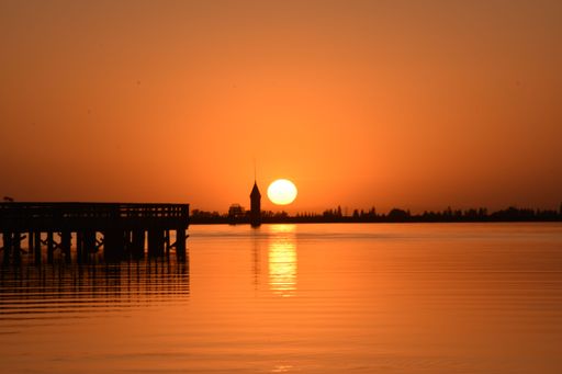 A serene sunset over a calm lake with a pier and a distant tower silhouette
