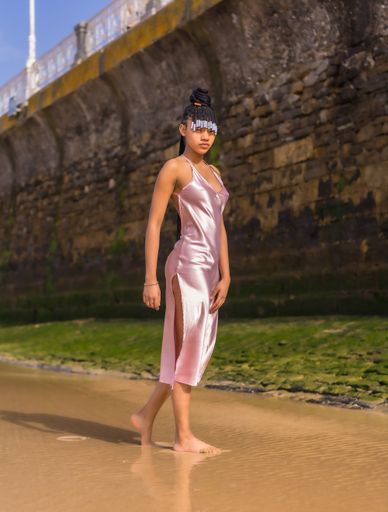 A dark-skinned American female with braids in a pink silky dress posing barefoot on the beach