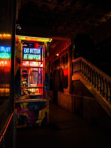 A neon-lit food stall in a dimly lit urban alley with colorful reflections