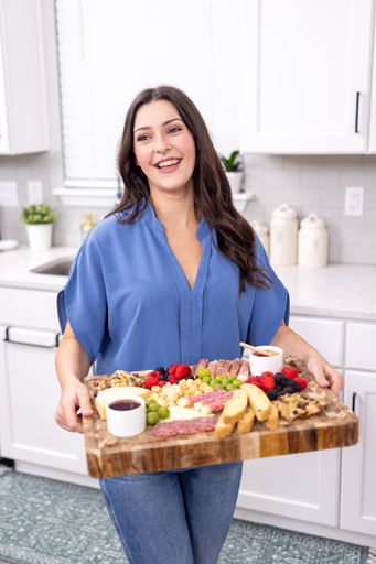 A brunette young female smiling in blue shirt in kitchen with charcuterie cheese board
