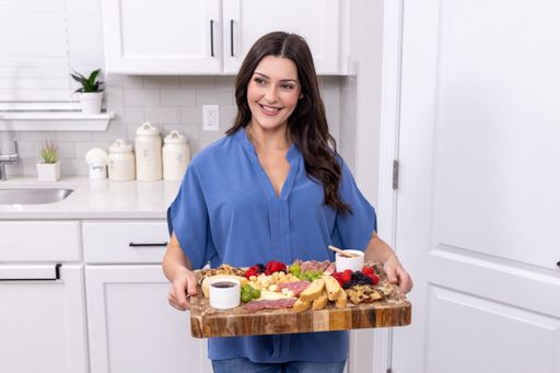 A brunette young female smiling in blue shirt in kitchen with charcuterie cheese board