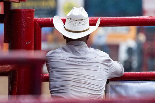 A man wearing a cowboy hat and shirt leaning on a red fence at a rodeo event.