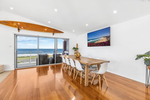 A modern dining room with ocean view, wooden floors, and surfboard decor on the wall.