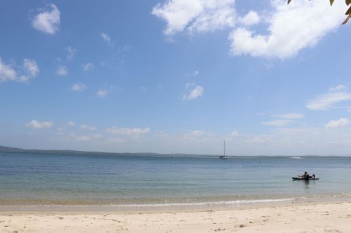 A serene beach scene with a clear blue sky, calm waters, and a person kayaking in the distance
