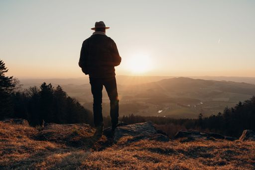 A Silhouette of a Middle Eastern man in a hat standing on a hilltop watching the sunset over a scenic valley in hiking area Bavarian Forest in Germany