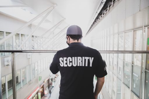 a male security guard with a black t-shirt and cap standing inside the building with big windows