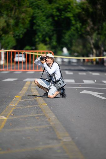 A male photographer in casual attire kneeling on a street taking photos with a camera on a sunny day.
