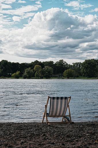A solitary beach chair facing a serene river under a partly cloudy sky.