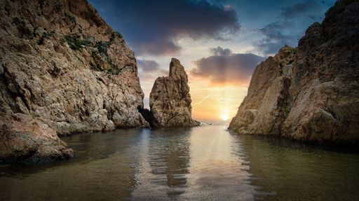 A sunset view between rocky cliffs with a calm sea reflecting the colorful sky