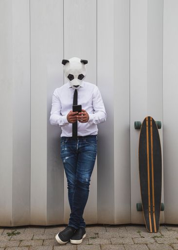 A male in a white shirt, tie, and panda head mask chatting with a smartphone, leaning on the wall