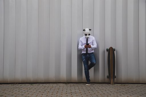 A male in a white shirt, tie, and panda head mask chatting with a smartphone, leaning on the wall