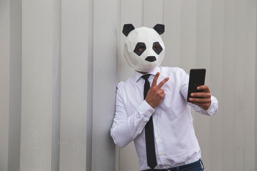 A male in a white shirt, tie, and panda head mask taking smartphone selfie against the gray wall