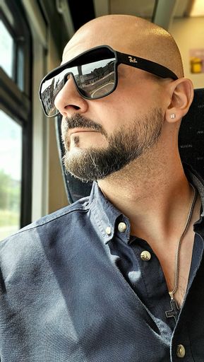 A bearded man wearing sunglasses and a cross necklace, looking out the window of a train