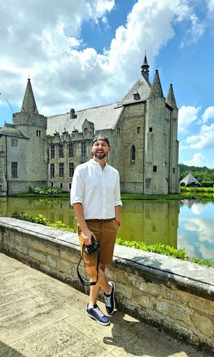 A Man holding a camera standing in front of a historic castle with a moat, under a bright blue sky.