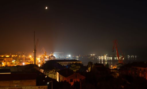 The night view of a harbor with illuminated cranes and buildings under a crescent moon. Pula, Croatia