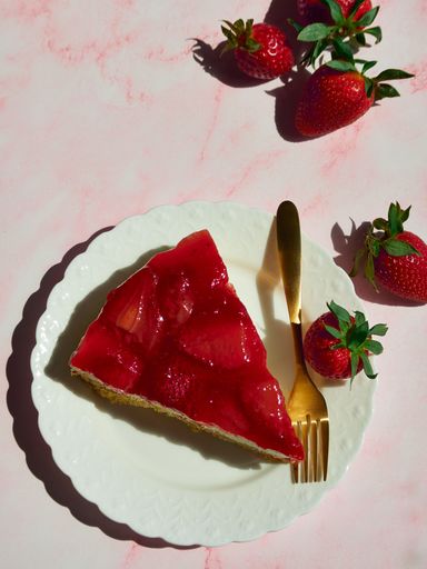 A slice of strawberry cheesecake on a white plate with a golden fork and fresh strawberries on a pink background