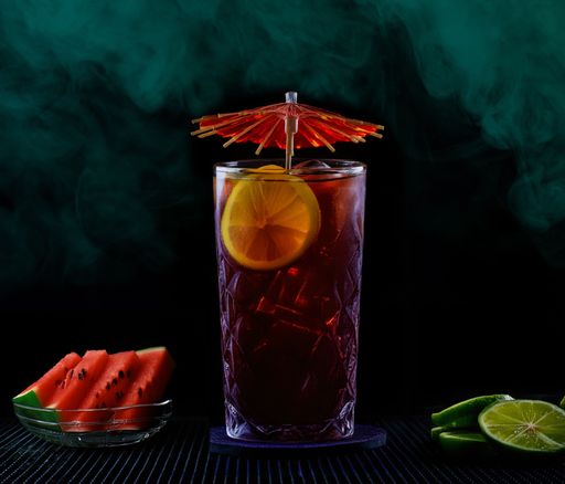 A refreshing glass of iced tea with lemon slices and a cocktail umbrella, accompanied by watermelon slices and lime on a dark background