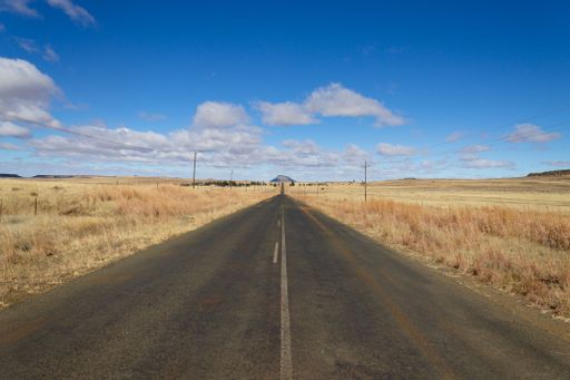 A long straight road through a dry rural landscape under a blue sky with scattered clouds.