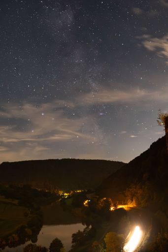 A stunning night sky with the milky way over a serene landscape with a river and hills illuminated by warm lights.