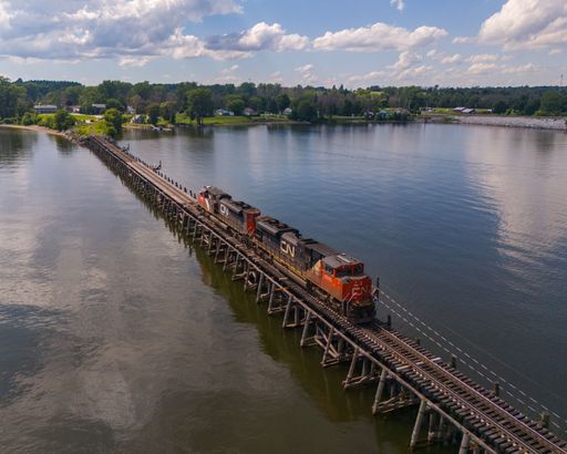 An aerial shot over a long train crosses a wooden trestle bridge over a calm lake under a blue sky with white clouds