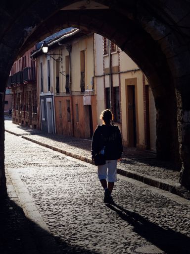 A vertical back shot of a person walking through a cobblestone street under an archway in a historic European town on a sunny day