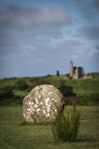 Ancient standing stone in a grassy field with ruins in the background under a blue sky, Cornwall, England