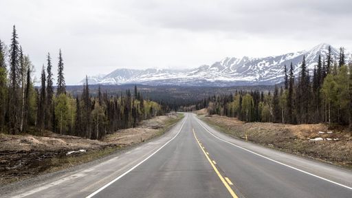 An empty long highway through pine forest with snowy mountains in the background in winter
