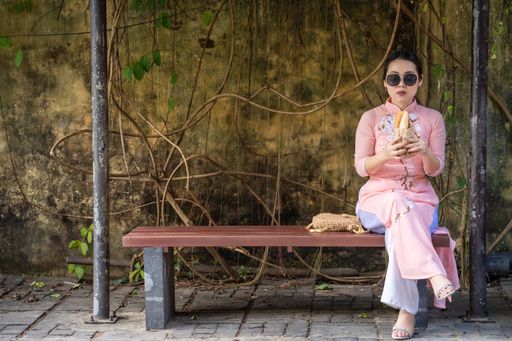 A Vietnamese woman in pink ao dai sitting on a bench at a park while eating bread